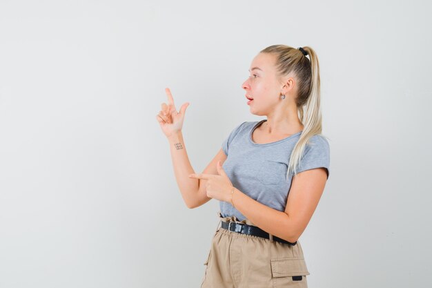 Jeune femme pointant vers le haut et de côté en t-shirt, pantalon et à la surprise. vue de face.