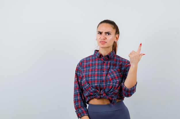 Jeune femme pointant vers le haut dans une chemise à carreaux, un pantalon et l'air sérieux, vue de face.