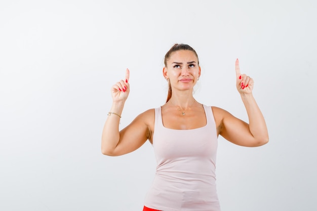 Jeune femme pointant vers le haut en débardeur beige et à l'insatisfaction, vue de face.