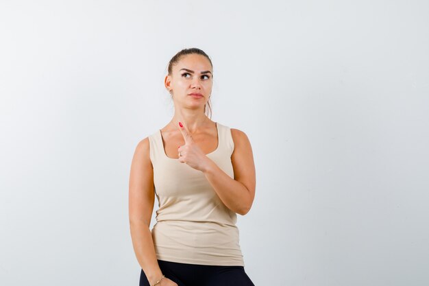 Jeune femme pointant vers le haut en débardeur et à la pensif. vue de face.