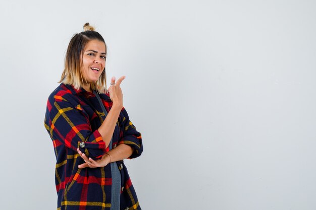 Jeune femme pointant vers le haut, debout sur le côté en chemise à carreaux décontractée et l'air heureuse.