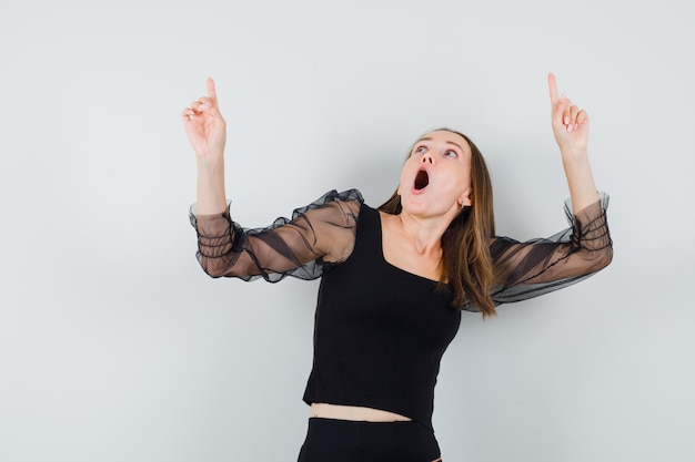 Jeune femme pointant vers le haut avec les deux index en chemisier noir et pantalon noir et à la surprise. vue de face.