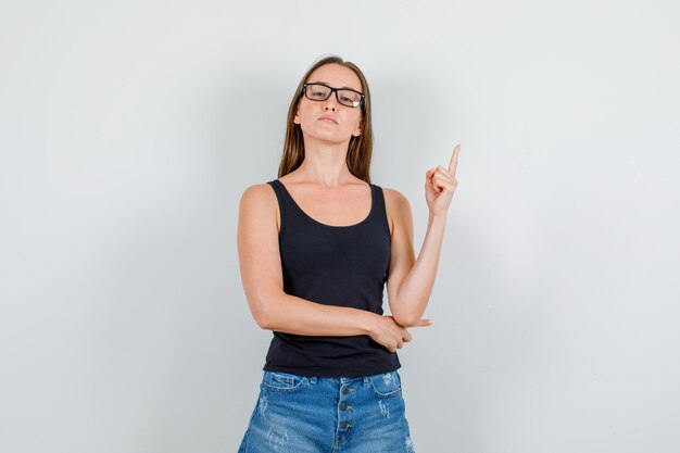 Jeune femme pointant vers le haut du doigt en maillot, shorts, lunettes et à la confiance