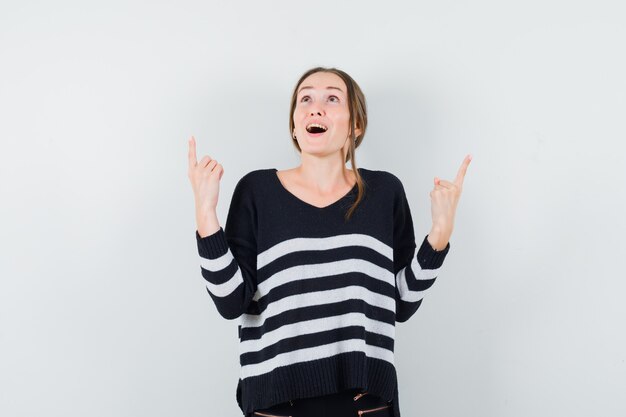 Photo gratuite jeune femme pointant vers le haut avec l'index en maille rayée et pantalon noir et à la surprise