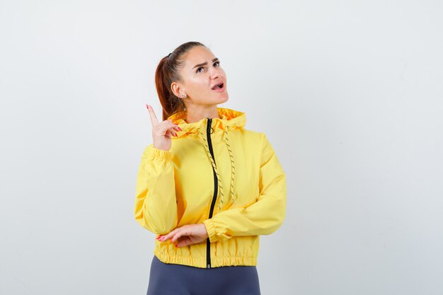 Jeune femme pointant vers le haut, ouvrant la bouche en veste jaune et l'air étonné. vue de face.