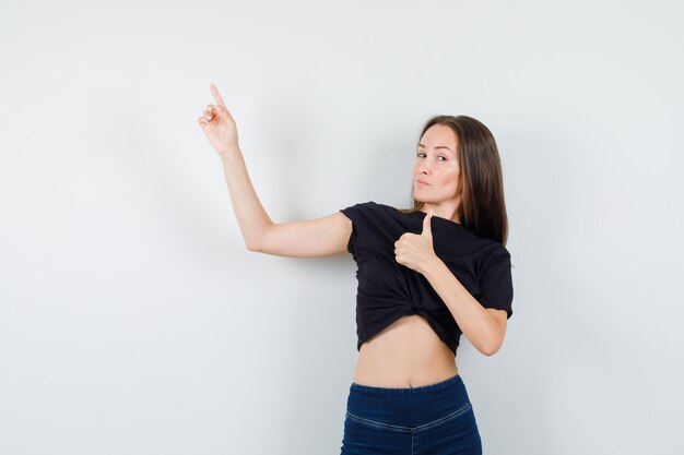 Jeune femme pointant vers le haut avec le pouce vers le haut en chemisier noir, pantalon et à la confiance.