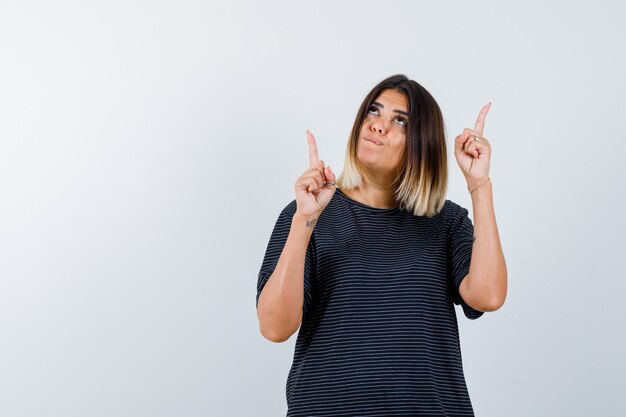 Jeune femme pointant vers le haut en robe de polo et à la vue réfléchie, de face.