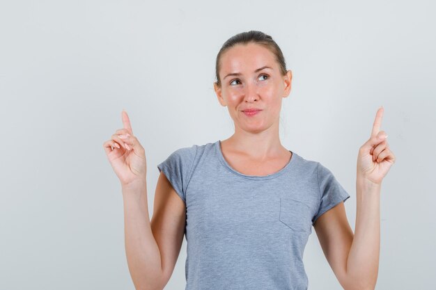 Jeune femme pointant vers le haut en t-shirt gris et à la rêveuse, vue de face.