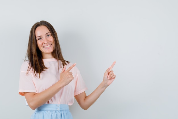 Jeune femme pointant vers le haut en t-shirt, jupe et à la vue de face, heureux.