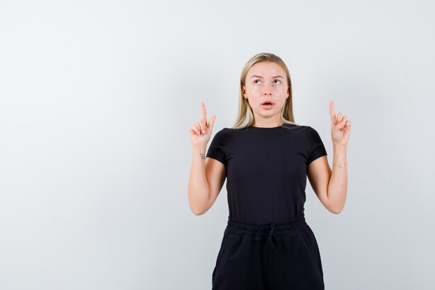 Jeune femme pointant vers le haut en t-shirt, pantalon et à la vue réfléchie, de face.