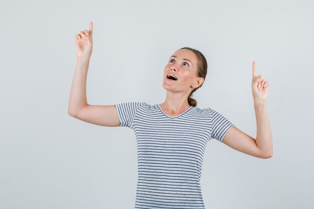 Jeune femme pointant vers le haut en t-shirt et regardant étonné, vue de face.