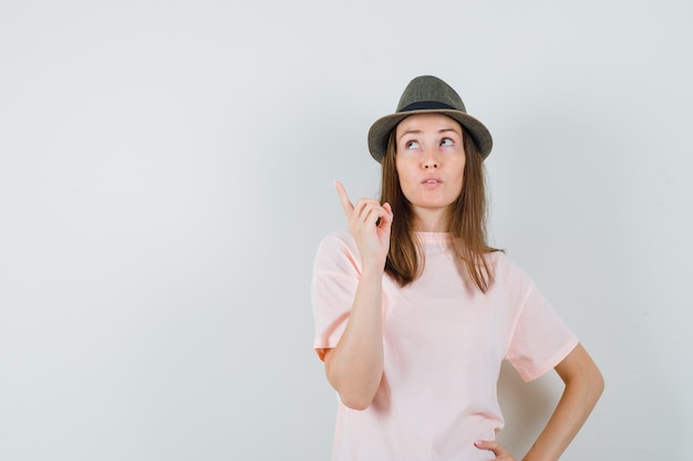Jeune femme pointant vers le haut en t-shirt rose, chapeau et à la recherche concentrée. vue de face.