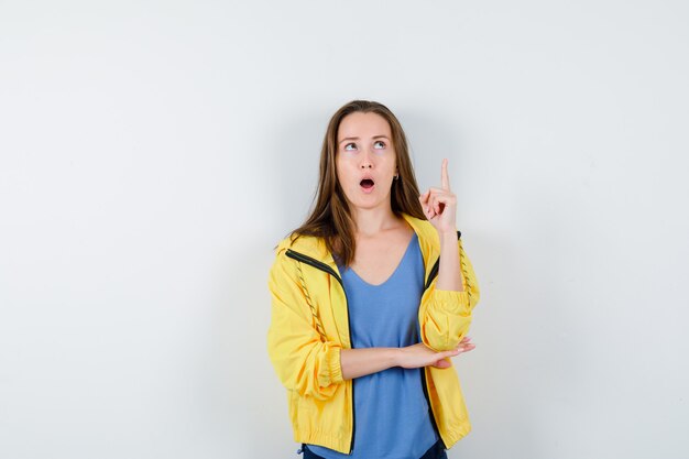 Jeune femme pointant vers le haut en t-shirt, veste et l'air surpris, vue de face.