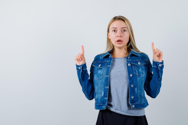 Jeune femme pointant vers le haut en t-shirt, veste en jean, jupe et l'air étonné