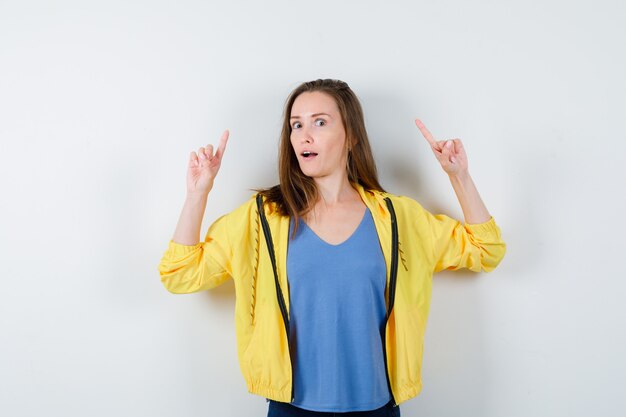 Jeune femme pointant vers le haut en t-shirt, veste et semblant curieuse, vue de face.