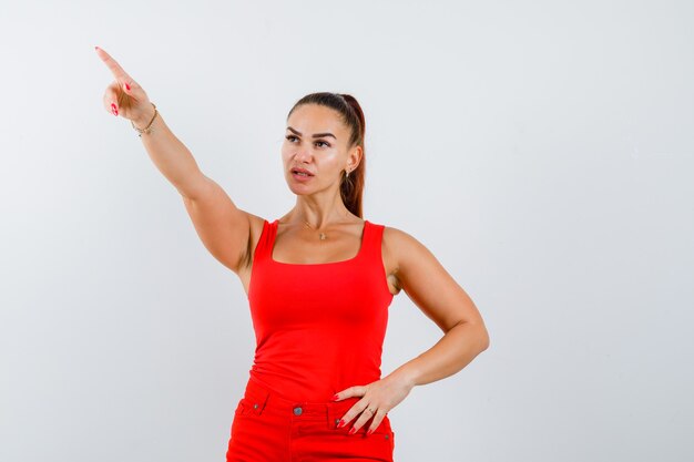 Jeune femme pointant vers le haut, tenant la main sur la taille en débardeur rouge, pantalon et à la nostalgie, vue de face.