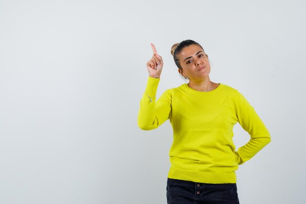 Jeune femme pointant vers le haut et tenant la main sur la taille en pull jaune et pantalon noir et l'air heureux