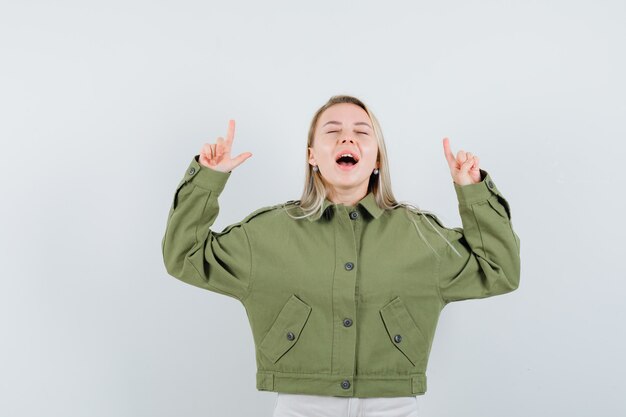 Jeune femme pointant vers le haut tout en criant en veste verte, jeans, vue de face.