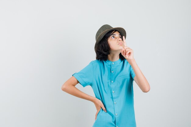 Jeune femme pointant vers le haut tout en écoutant en chemise bleue, chapeau et à la recherche concentrée.