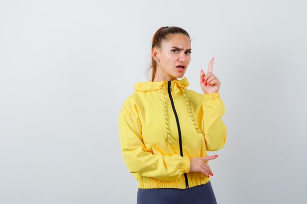 Jeune femme pointant vers le haut en veste jaune et semblant anxieuse. vue de face.