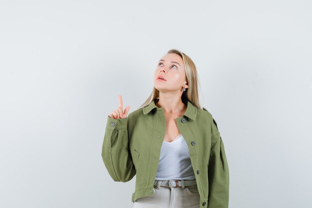 Jeune femme pointant vers le haut en veste, pantalon et à la recherche concentrée. vue de face.