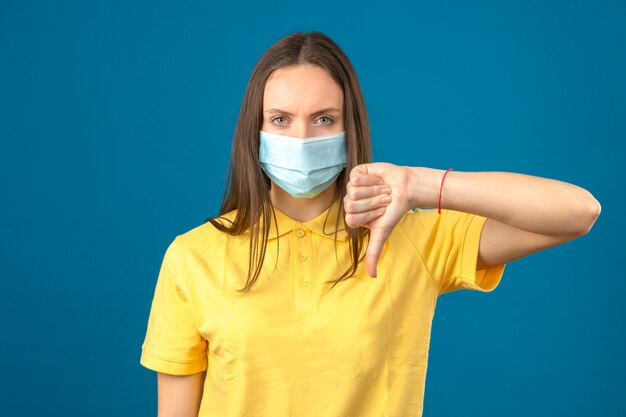 Jeune femme en polo jaune et masque de protection médicale faisant signe de pouce vers le bas regardant sérieusement la caméra sur fond bleu isolé