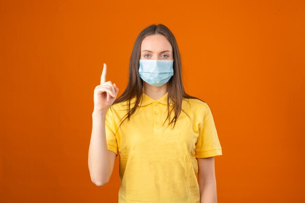 Jeune femme en polo jaune et masque de protection médicale regardant le doigt pointé vers le haut sur fond orange