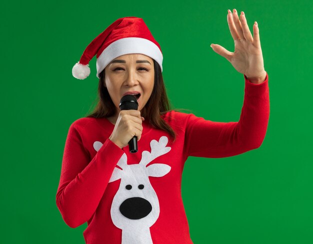 Jeune femme portant bonnet de noel de Noël et pull rouge tenant le microphone chantant heureux et sorti debout sur fond vert