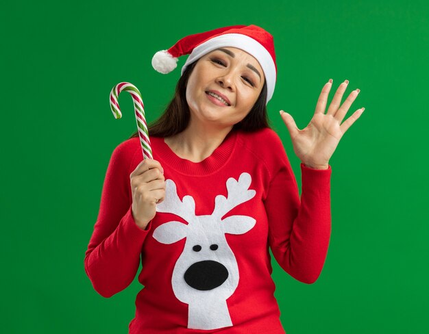 Jeune femme portant chapeau de Père Noël et pull rouge tenant la canne en bonbon regardant la caméra heureux et joyeux en agitant avec la main debout sur fond vert