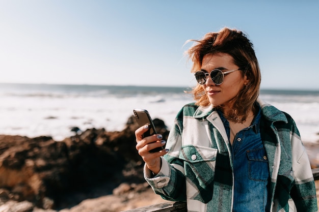 Jeune femme portant une chemise en jean avec des lunettes de soleil posant sur la plage tout en regardant le smartphone
