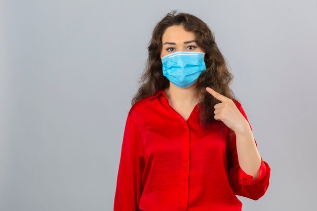 Jeune femme portant un chemisier rouge dans un masque de protection médicale regardant la caméra et pointant le doigt pour masquer le visage sur fond blanc isolé