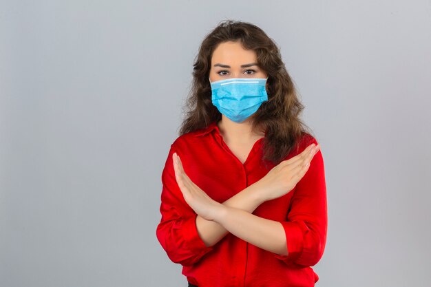 Jeune femme portant un chemisier rouge en masque de protection médicale debout avec les bras croisés sur la poitrine faisant le geste d'arrêt sur fond blanc isolé