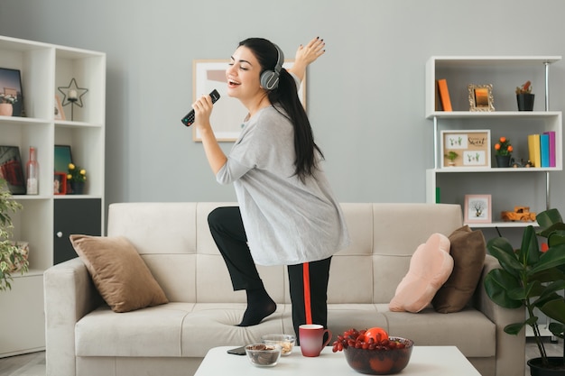 Jeune femme portant des écouteurs tenant une télécommande de télévision chante debout sur un canapé derrière une table basse dans le salon