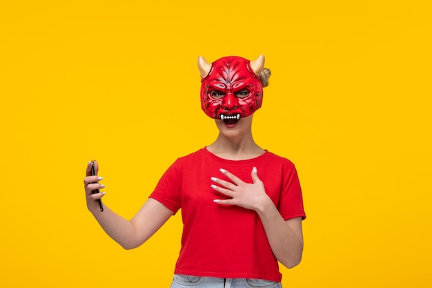Jeune femme portant un masque de diable effrayant à l'aide de son téléphone fond jaune moche