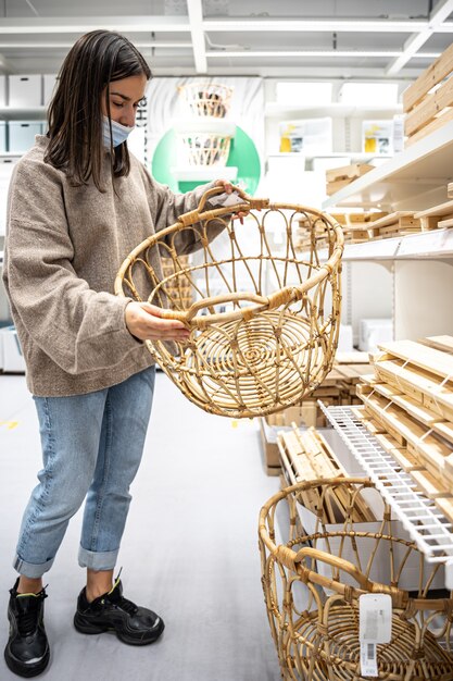 Une jeune femme portant un masque médical de protection choisit un panier dans un magasin avec des articles ménagers, faisant ses courses pendant la pandémie de coronavirus.