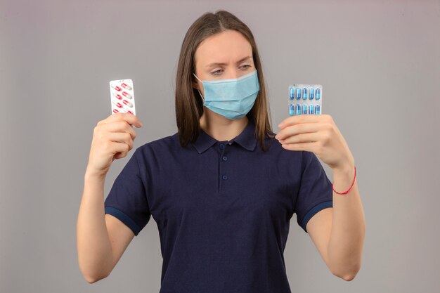 Jeune femme portant un polo bleu en masque médical confus en regardant blister avec des pilules dans les mains et en pensant faire le choix debout sur fond gris clair