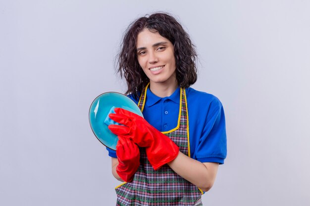 Jeune femme portant un tablier et des gants en caoutchouc plaque de lavage regardant la caméra avec le sourire sur le visage debout sur fond blanc