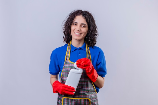 Jeune femme portant un tablier et des gants en caoutchouc tenant une bouteille avec des produits de nettoyage souriant joyeusement regardant la caméra debout sur fond blanc