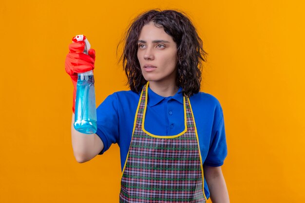 Jeune femme portant un tablier et des gants en caoutchouc tenant un spray de nettoyage à côté avec un visage sérieux, prêt à nettoyer sur mur orange