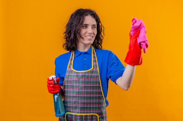 Jeune femme portant un tablier et des gants en caoutchouc tenant un spray de nettoyage et un tapis souriant avec visage heureux prêt yo nettoyer sur mur orange isolé
