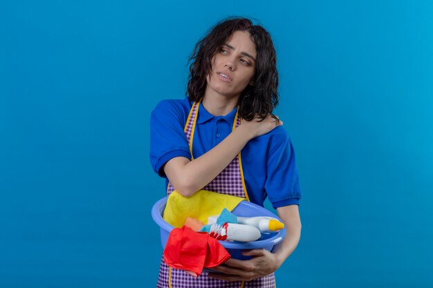 Jeune femme portant un tablier tenant le bassin avec des outils de nettoyage à l'épaule de toucher surmené ayant de la douleur sur le mur bleu