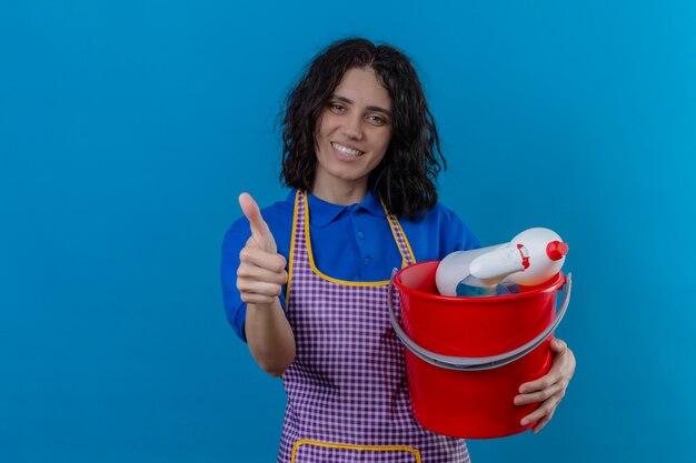 Jeune femme portant un tablier tenant un seau avec des outils de nettoyage souriant gai montrant les pouces vers le haut sur le mur bleu