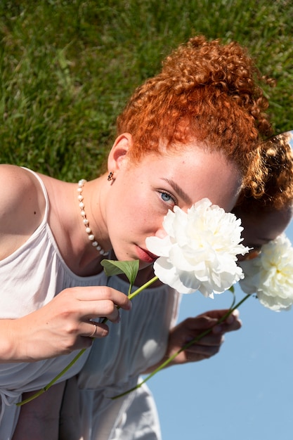 Jeune femme posant avec une fleur de pivoine dans le miroir sur l'herbe à l'extérieur