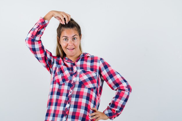 Jeune femme posant avec la main sur la tête en chemise à carreaux et à la drôle. vue de face.