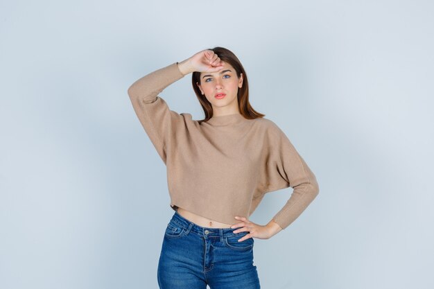 Jeune femme posant avec la main sur la tête dans un pull beige, un jean et l'air confiant. vue de face.