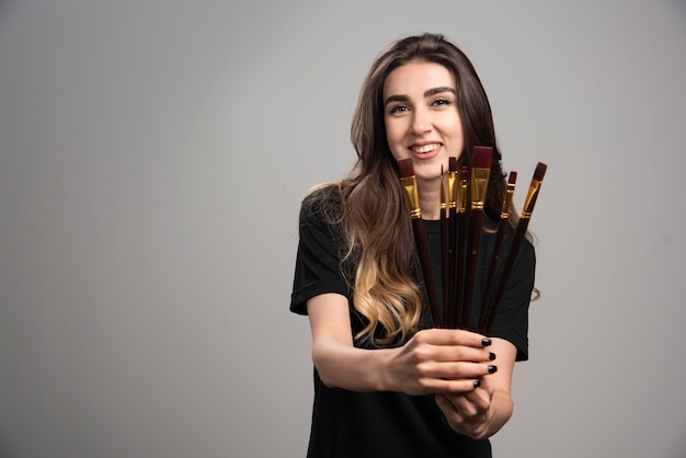 Jeune femme posant avec des pinceaux sur mur gris.