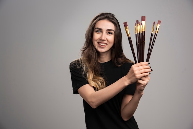 Jeune femme posant avec des pinceaux sur mur gris.