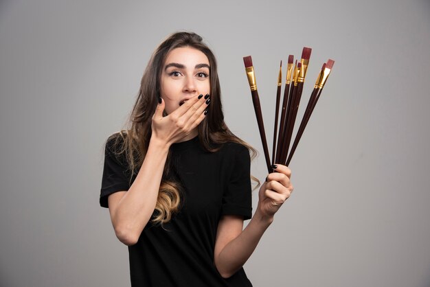 Jeune femme posant avec des pinceaux sur mur gris.