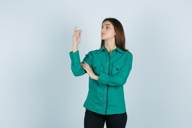 Jeune femme posant en pointant vers le haut en chemise verte, pantalon et magnifique. vue de face.