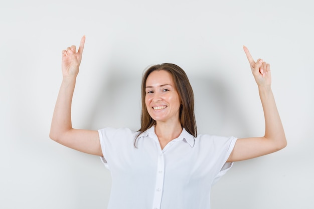 Photo gratuite jeune femme posant en pointant vers le haut en chemisier blanc et à la recherche de joie.
